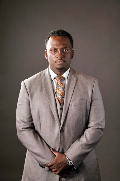 Man in a light gray suit with a patterned tie stands confidently against a plain background.
