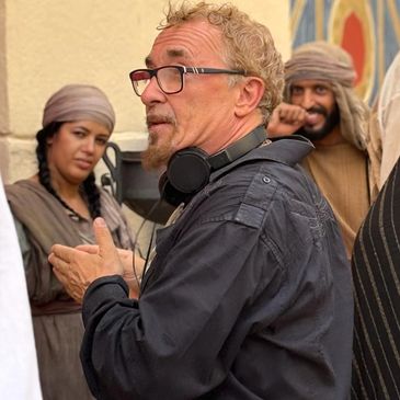 Man with glasses and headset surrounded by two people in traditional attire.