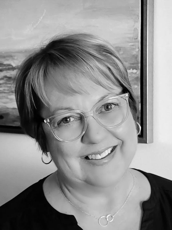 Smiling woman with short blonde hair and glasses in front of a painting.