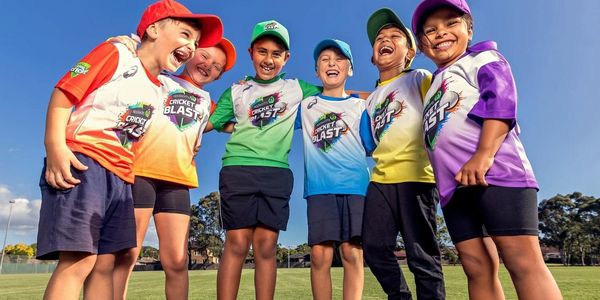 Group of kids in colorful cricket uniforms smiling on a field.