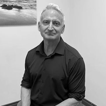 A mature man in a black shirt sitting indoors with a calm expression.