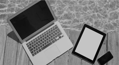 black and white image of a lap top, iPad, phone near a pool