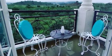 Cozy balcony setup with white iron chairs and a small table overlooking green hills.