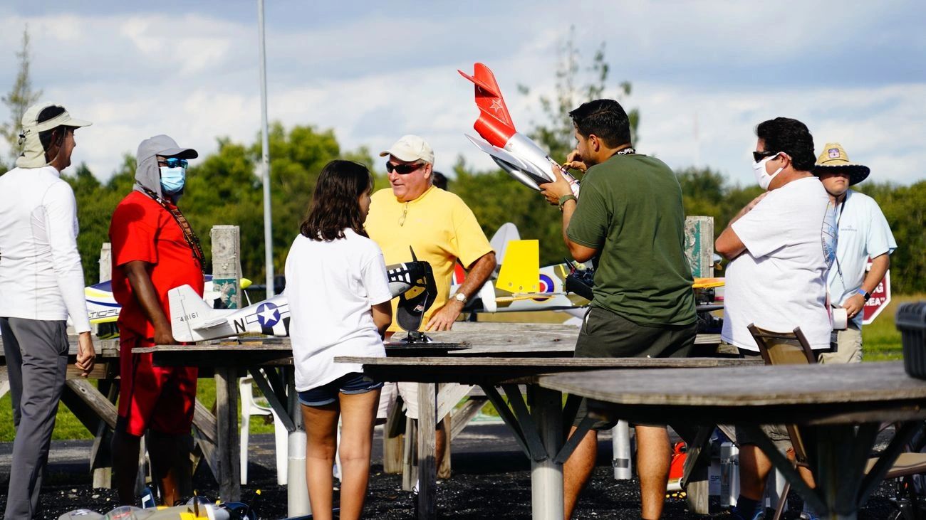 Group of people gathered around tables with model airplanes outdoors.