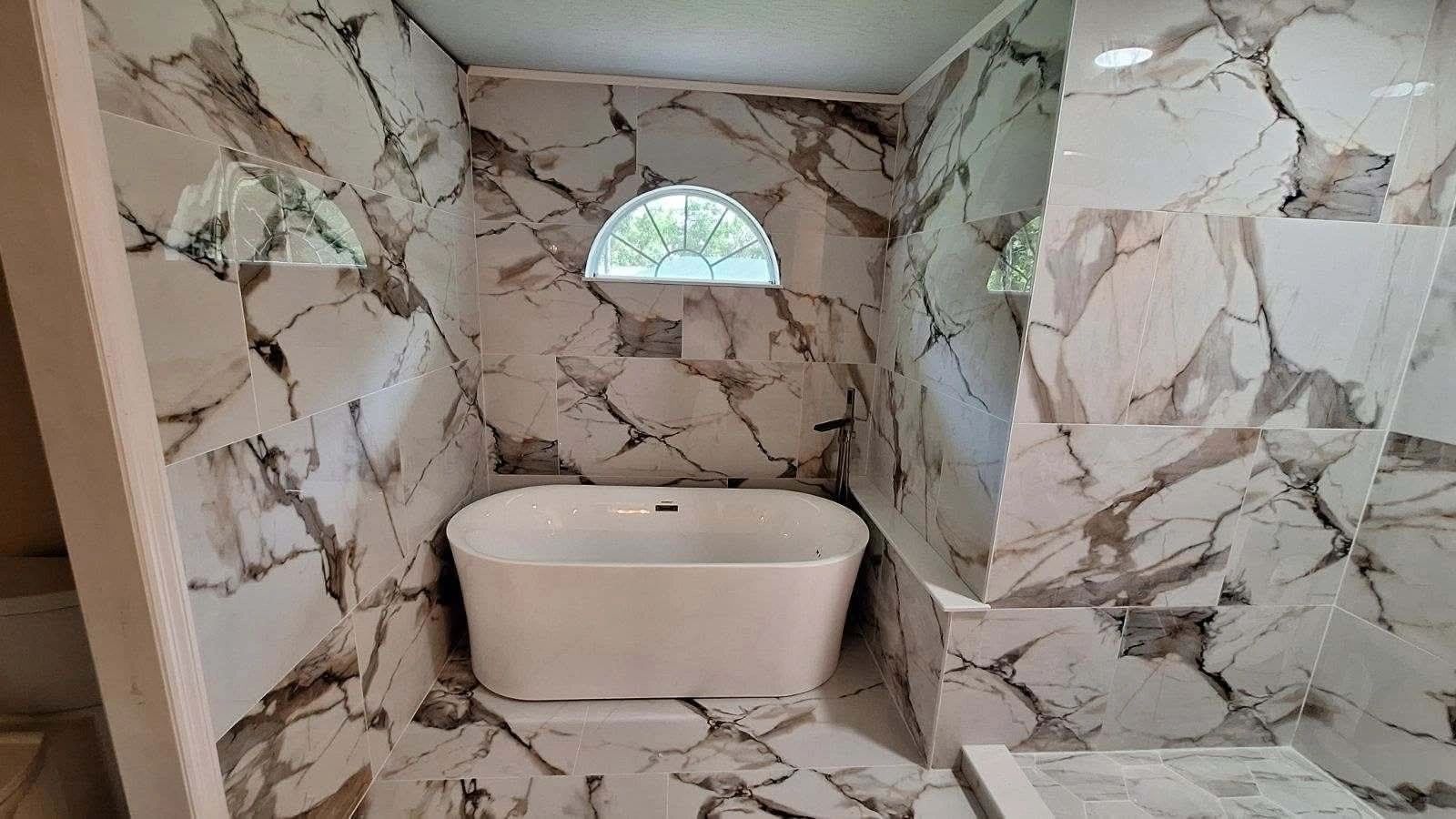 Modern bathroom with marble tiles and a standalone white bathtub under a half-moon window.