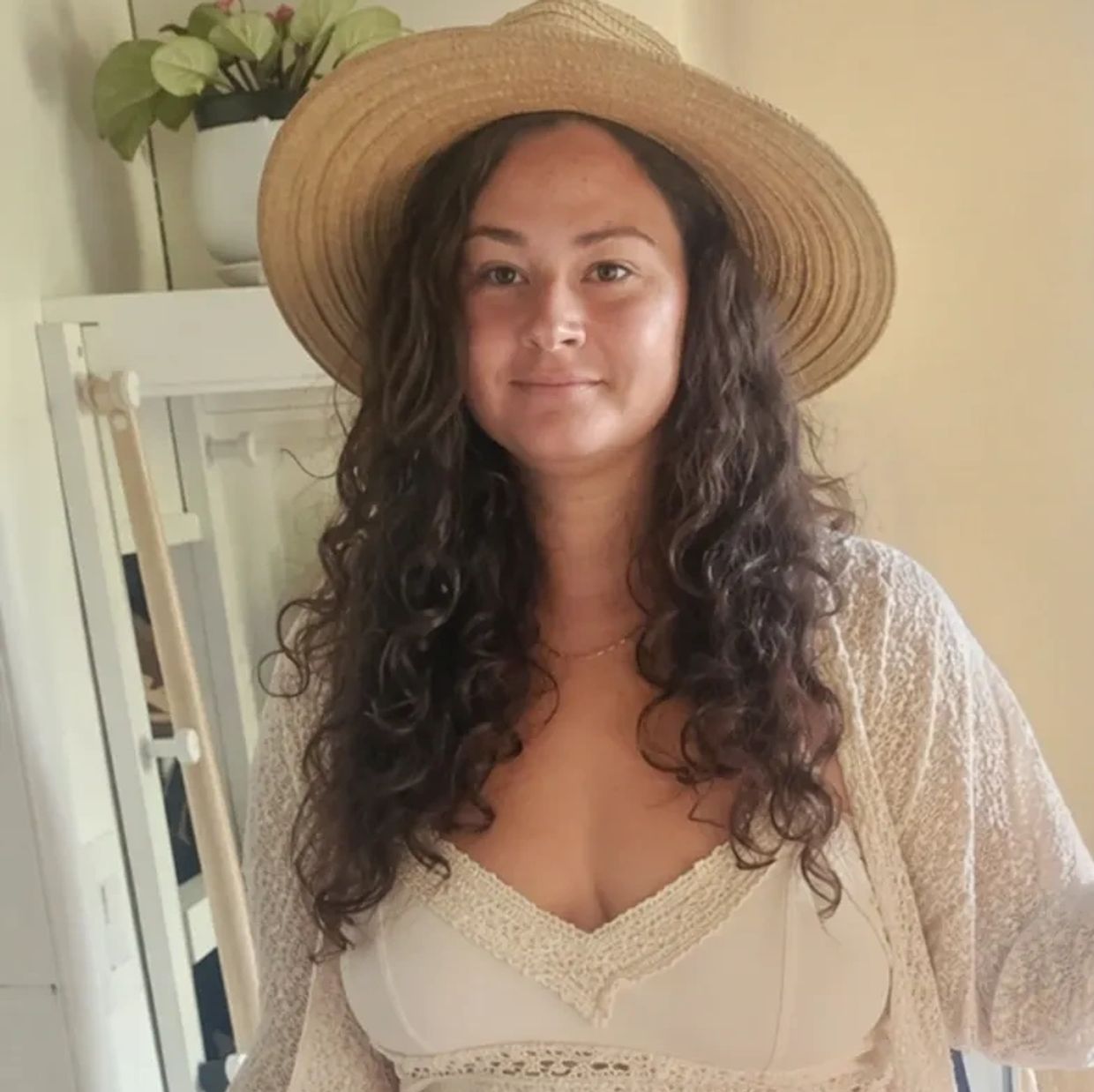 Woman with curly hair wearing a straw hat and white lace top.