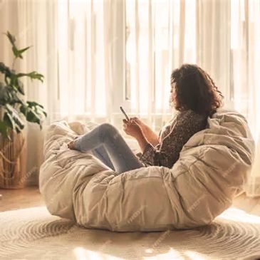 Woman relaxing on a cozy bean bag chair, using her phone in a sunlit room.