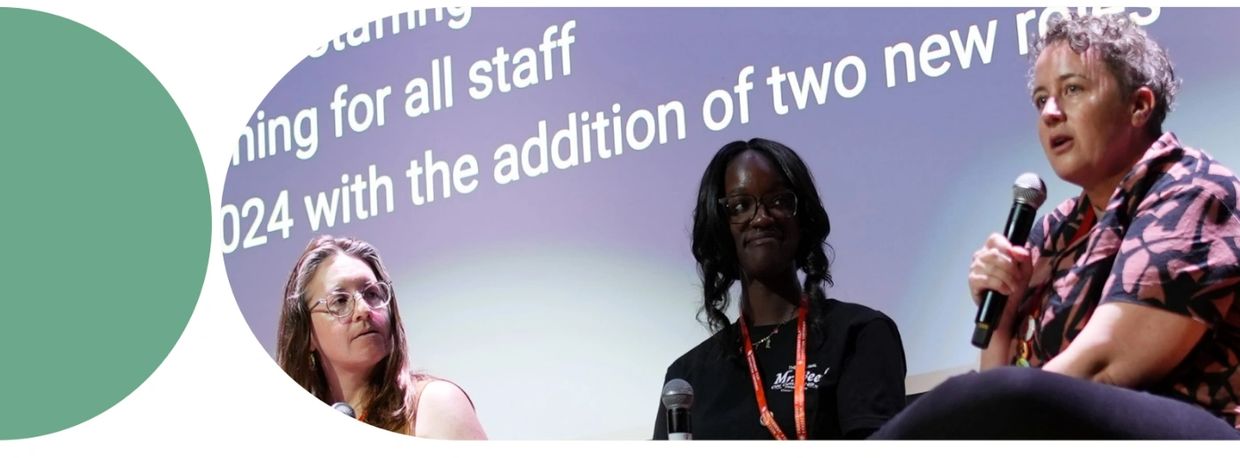 Three women sitting on a stage holding microphones, with a screen with large text captions behind th