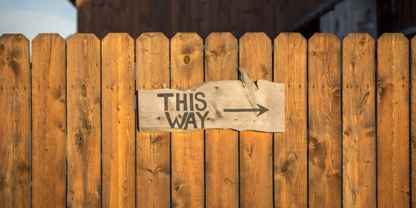 A wooden fence with a handwritten sign that says "this way" and an arrow pointing right.
