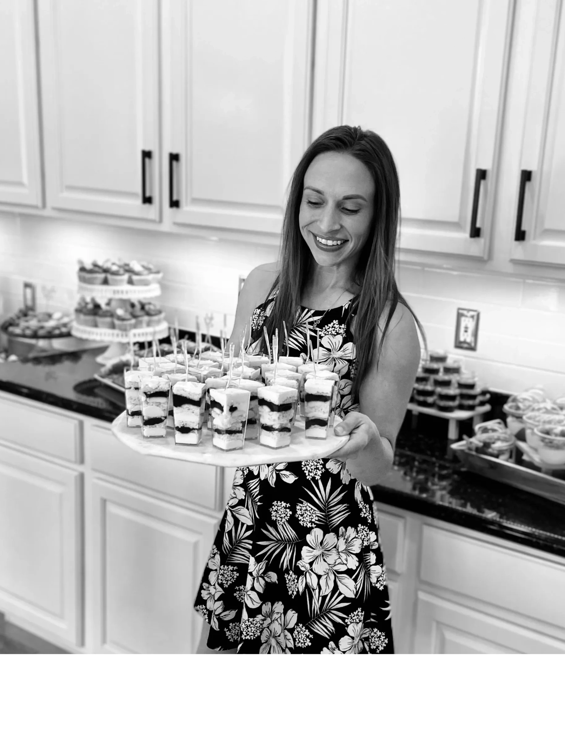 Smiling woman holding tray of layered dessert cups in a kitchen.