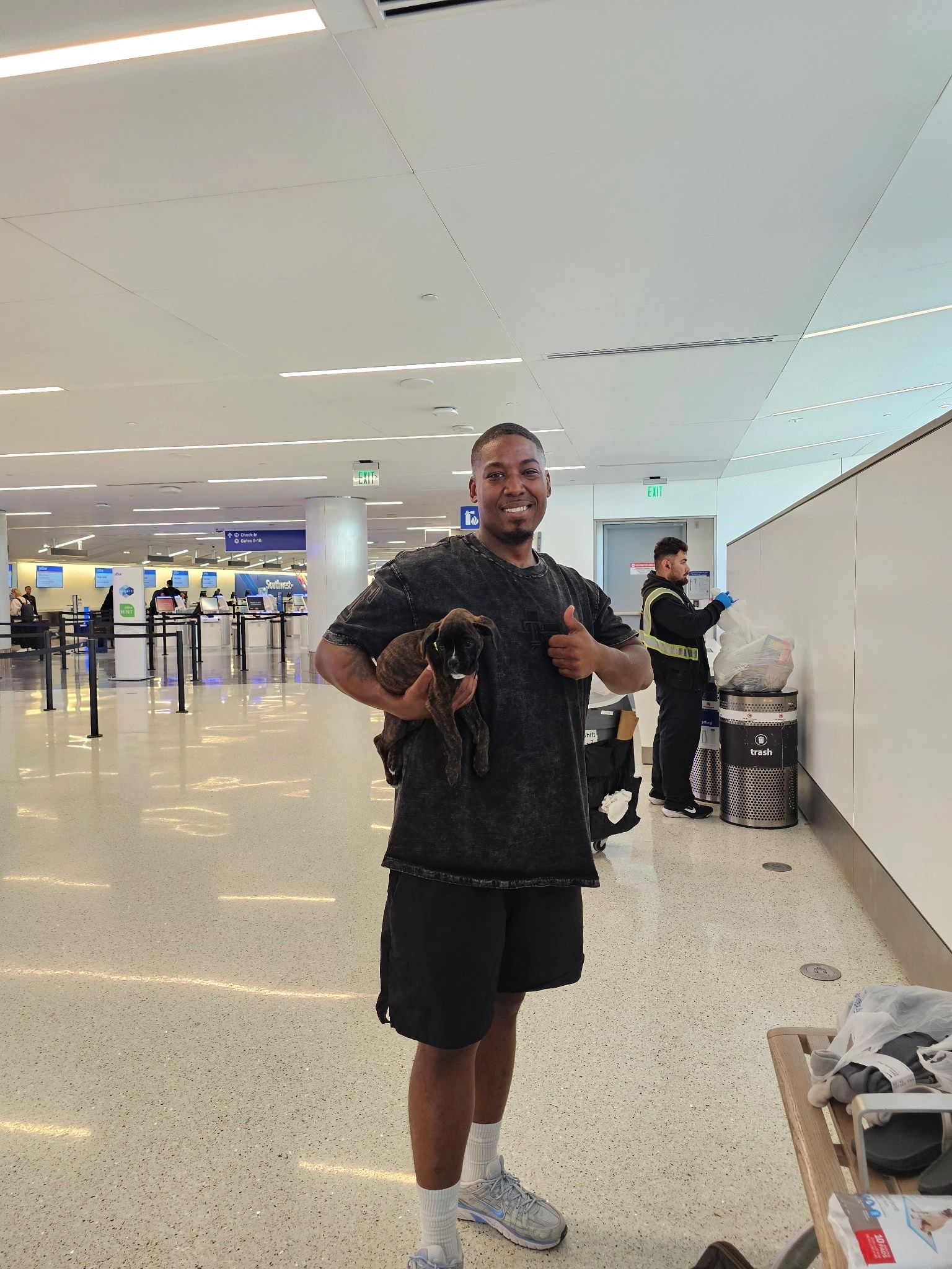 Man holding a puppy at an airport, giving thumbs up.