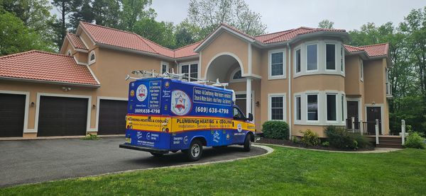 A plumbing service van parked outside a large suburban house with a red tile roof.