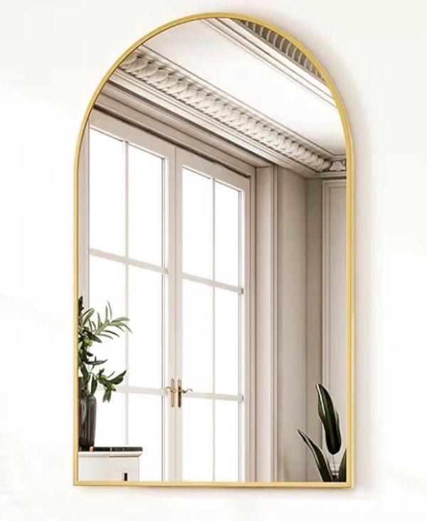 Elegant arched mirror with gold frame reflecting a bright window and indoor plants.