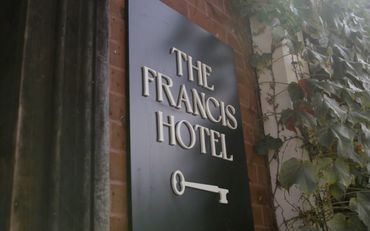 A sign for The Francis Hotel with a key symbol, mounted on a brick wall with ivy.