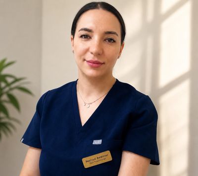 A smiling woman in navy blue scrubs with a name tag, standing indoors near a plant.