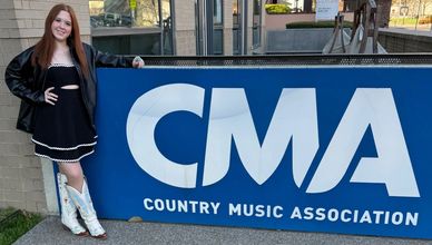 A woman in a black dress and white boots stands beside a CMA banner.