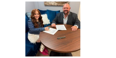 Two people signing documents at a round wooden table in an office.