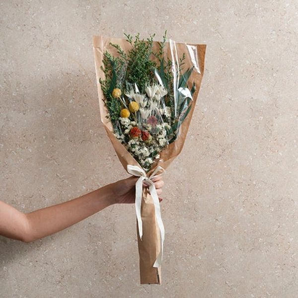 Hand holding a rustic bouquet of dried flowers wrapped in brown paper and plastic.
