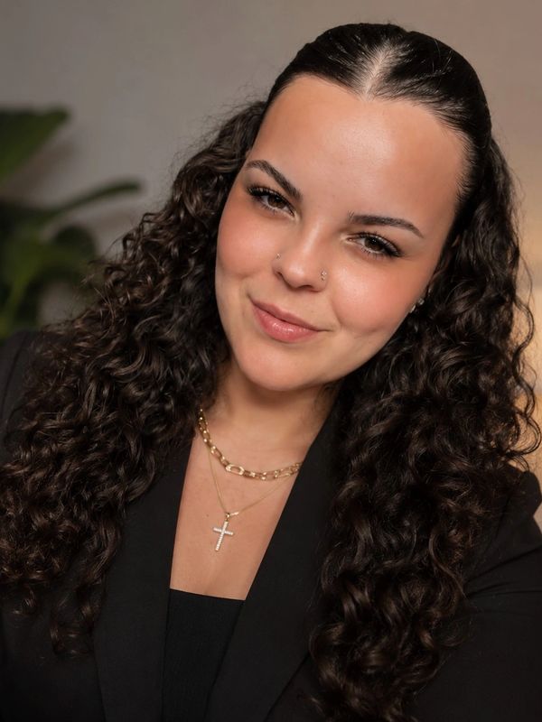 Woman with curly hair smiling, wearing a black blazer and a cross necklace.