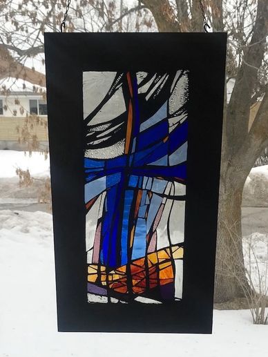 Framed stained glass art suspended in a window. Blue, yellow, amber, burnt orange, purple, and clear