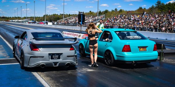 Two cars line up for a drag race with a woman standing between them on the track.