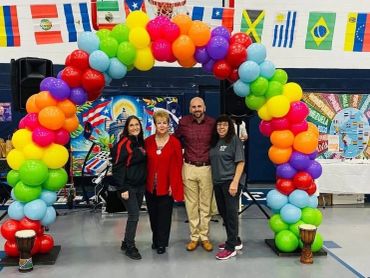 Four people standing under a colorful balloon arch in a gym.