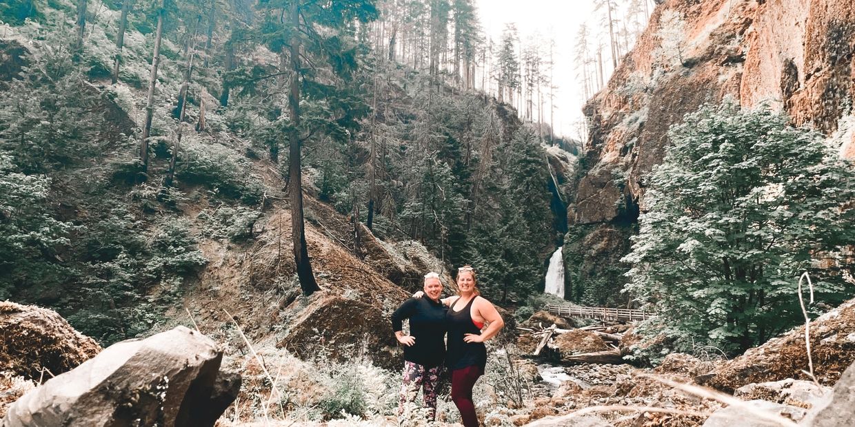 Two women posing in front of a waterfall in a forested canyon.