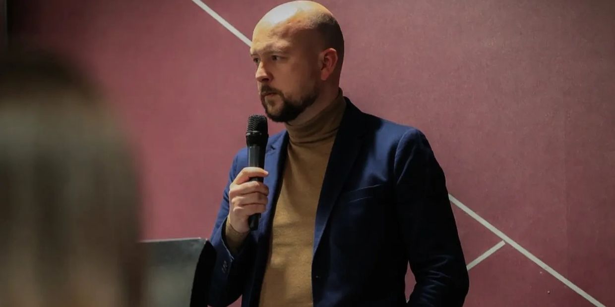 A man in a blazer and turtleneck speaking into a microphone.