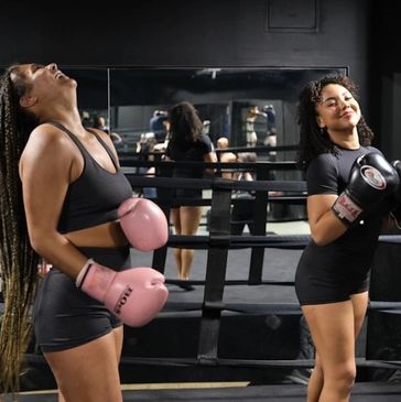 Two women in boxing gear smiling and enjoying their time in a boxing ring.