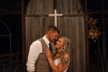 A couple lovingly embraces in front of a wooden door with a cross at night.