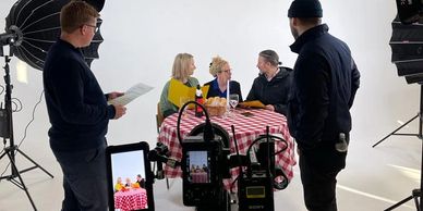 Film crew captures a group at a table with red checkered cloth in a studio.