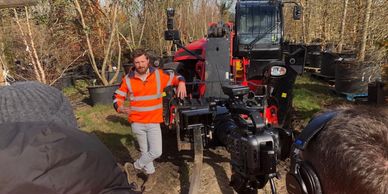 Man in orange safety vest filmed with camera near red machinery outdoors.