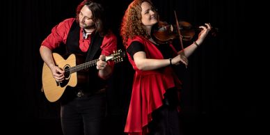 A man and woman joyfully playing guitar and violin on stage.