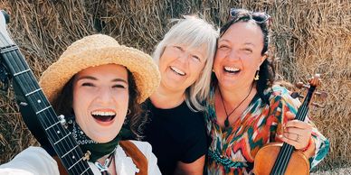 Three women smiling with musical instruments in front of hay bales.