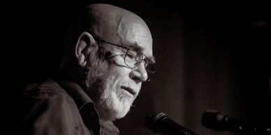 Elderly man with glasses speaking into a microphone in a dim setting.