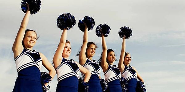 Five cheerleaders in blue and white uniforms raising pom-poms against a cloudy sky.