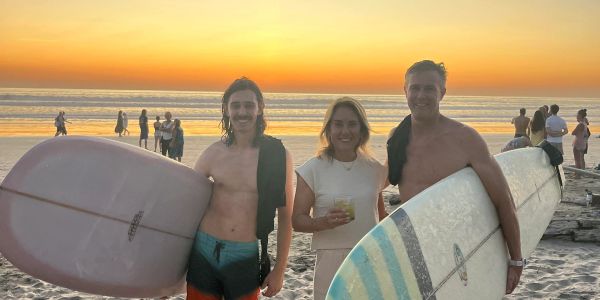 Three friends on the beach at sunset, two holding surfboards.