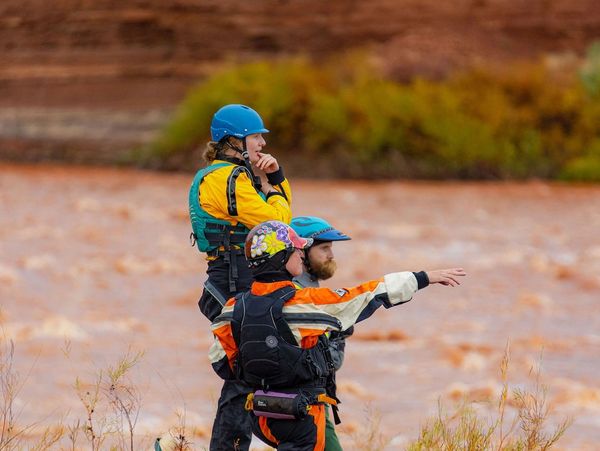 Three people in helmets and life jackets near a river, one pointing ahead.