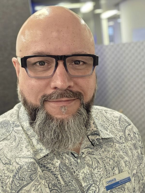 Smiling man with glasses and a patterned shirt wearing a name tag.