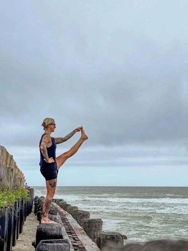 A tattooed woman stretches her leg on a wooden post by the ocean.