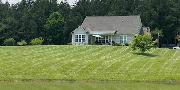 A house with a large, neatly mowed lawn by a pond and surrounded by trees.