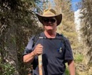 Man wearing a cowboy hat and sunglasses holding a walking stick in a forest.