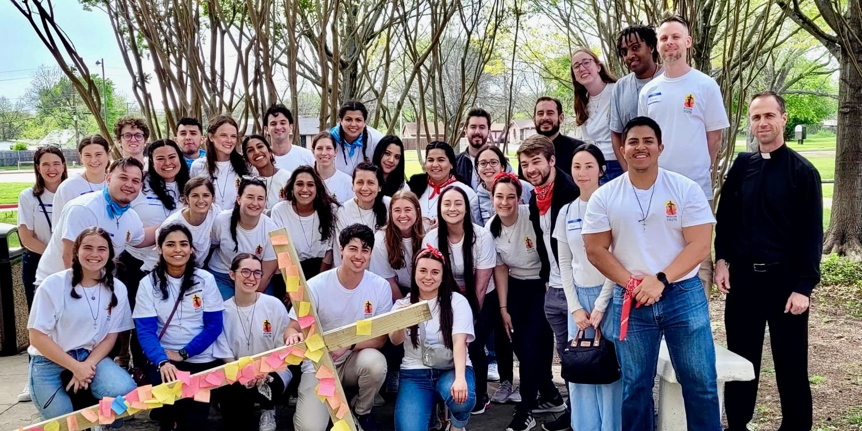 Large group of smiling young people with a priest in a park.