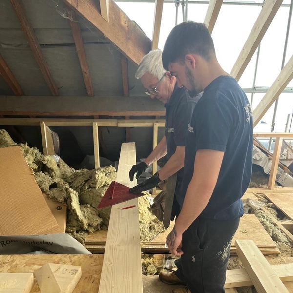Two men measuring wood in an attic under construction.