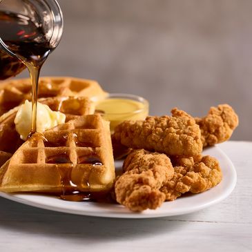 Syrup pouring over waffles with fried chicken on a white plate.