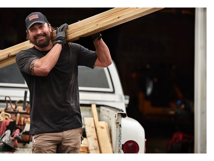 Smiling worker carrying wooden planks over his shoulder near a truck.