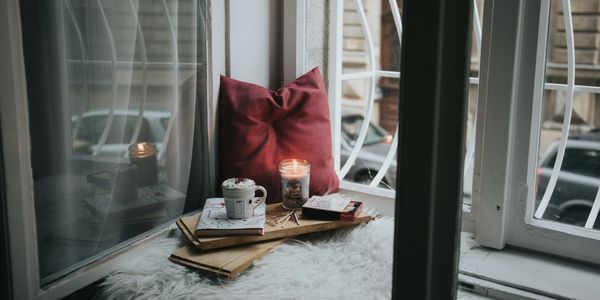 Cozy window nook with candle, hot drink, and red pillow on a fluffy white rug.