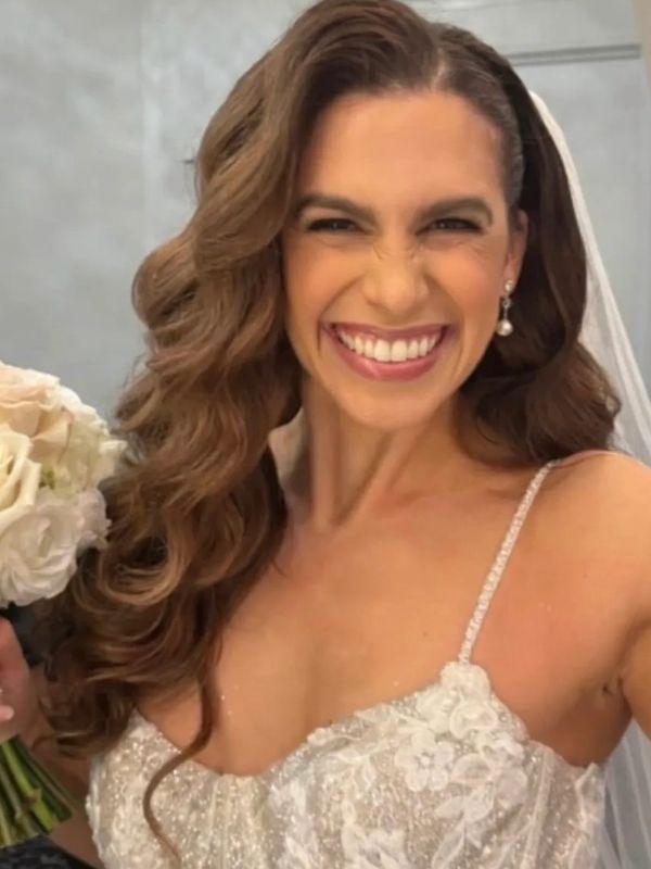 A joyful bride with long wavy hair smiles brightly, holding a bouquet of white flowers.