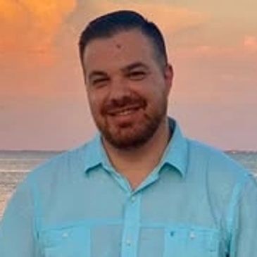 Man smiling outdoors with a sunset background.