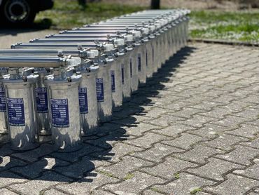 A long row of shiny metal containers with blue labels arranged on paved ground.